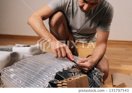 Man unboxing and cutting plastic air cushion wrap packaging with utility knife. Carefully preparing office chair components for assembly. Focusing on home improvement and diy furniture construction. 135797543