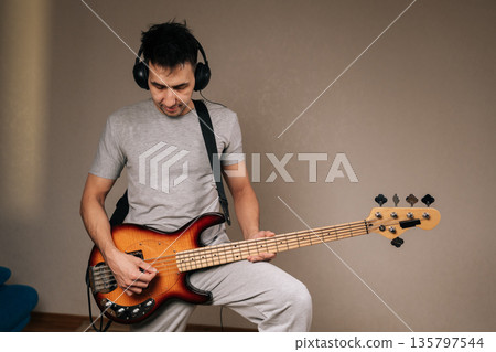 Portrait of young man wearing headphones diligently practicing bass guitar, focusing on strings to improve musical skills, engaging in passion and enjoying creative process indoors. 135797544