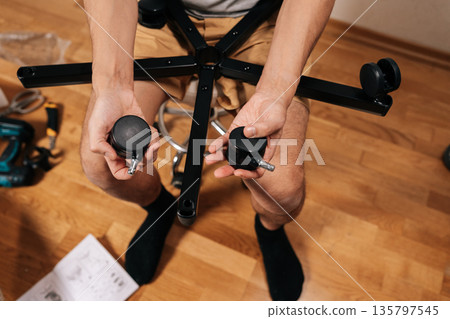 Top view of man assembling new ergonomic office chair, inserting wheel castor into the chair base, focusing on construction and diy furniture repair at home. 135797545