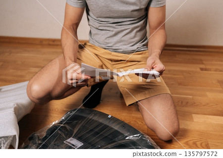 Cropped shot of man following written instructions to assemble new flat-pack furniture, focusing on steps to build office chair on te hardwood floor, common home improvement and diy task. Cropped shot of man following written instructions to assemble new flat-pack furniture, focusing on steps to build office chair on te hardwood floor, common home improvement and diy task. 135797572