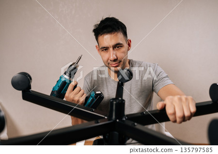 Portrait of contractor male assembling new office chair indoors, using electric drill to secure parts, focused diy furniture project showing tools, skill and home workspace setup. Portrait of contractor male assembling new office chair indoors, using electric drill to secure parts, focused diy furniture project showing tools, skill and home workspace setup. 135797585