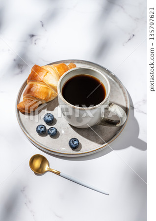 Freshly brewed black coffee in a cup on a plate on a white marble background with croissants Freshly brewed black coffee in a cup on a plate on a white marble background with croissants 135797621