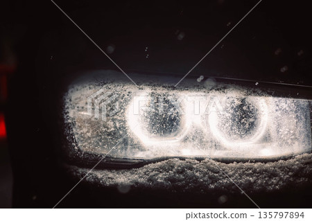A car headlight covered in snow at night. Winter driving at nighttime. Heavy snow, blizzard, storm weather. Automobile LED light in the darkness. 135797894