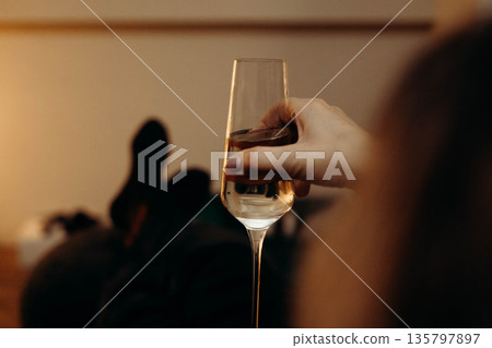 A hand holding a flute glass filled with sparkling wine or champagne. The image focuses on the drink and glassware against a warmly lit interior 135797897