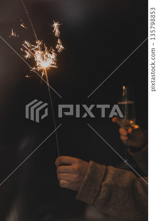A person holding a burning sparkler and a glass of champagne in a dark, cozy atmosphere. Festive New Year or Christmas celebration, holiday magic A person holding a burning sparkler and a glass of champagne in a dark, cozy atmosphere. Festive New Year or Christmas celebration, holiday magic 135798853