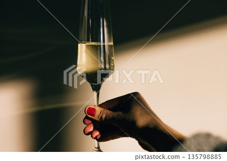 Female hand holding champagne glass with red manicure in dramatic light and shadow. Festive toast, celebration, holiday greeting, elegant lifestyle. 135798885