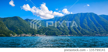 View from the side of Lake Como of the mountainous shores, and the settlements on the lake's shore. The water surface of the lake from a tourist boat. Tourist attractions in Italy. High quality photo 135799141