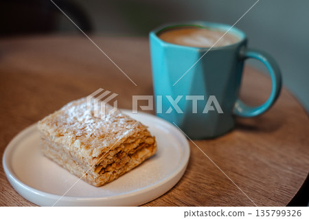 Delicate Napoleon cake slice on a white plate with a modern blue coffee mug of coffee with milk, cappuccino in a cozy cafe setting. Perfect for food blogs, menus, or aesthetic culinary content. Delicate Napoleon cake slice on a white plate with a modern blue coffee mug of coffee with milk, cappuccino in a cozy cafe setting. Perfect for food blogs, menus, or aesthetic culinary content. 135799326