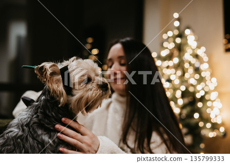 Festive Christmas moment at home - a young white woman sharing a cozy, joyful evening with a small dog Yorkshire terrier near a decorated Christmas tree, warm lights, hygge mood and holiday lifestyle. Festive Christmas moment at home - a young white woman sharing a cozy, joyful evening with a small dog Yorkshire terrier near a decorated Christmas tree, warm lights, hygge mood and holiday lifestyle. 135799333