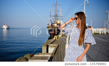 Young woman standing on pier and adjusting sunglasses by the sea Young woman standing on pier and adjusting sunglasses by the sea 135799991