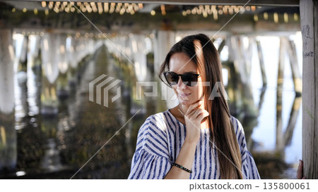Stylish woman in sunglasses under wooden pier on sunny day Stylish woman in sunglasses under wooden pier on sunny day 135800061