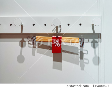 A modern wooden hanger with a red sale tag is displayed in a clean retail setting with strong shadows, perfect for themes of shopping, discounts, and seasonal retail marketing and shopping Store scene A modern wooden hanger with a red sale tag is displayed in a clean retail setting with strong shadows, perfect for themes of shopping, discounts, and seasonal retail marketing and shopping Store scene 135800825