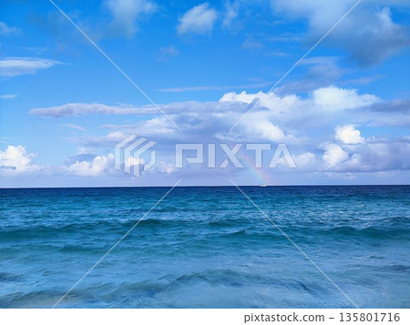Ocean view under a bright blue sky with scattered white clouds. A rainbow is visible on the horizon, adding a touch of color between the sea and sky Ocean view under a bright blue sky with scattered white clouds. A rainbow is visible on the horizon, adding a touch of color between the sea and sky 135801716