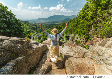 Young woman standing on rocky viewpoint with raised arms overlooking lush tropical valley and distant hills, freedom and adventure concept in summer travel landscape Young woman standing on rocky viewpoint with raised arms overlooking lush tropical valley and distant hills, freedom and adventure concept in summer travel landscape 135803399