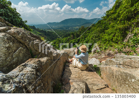 Young woman sitting on rocky ledge photographing lush tropical valley and distant hills, wide angle travel scene with green forest landscape, summer adventure and outdoor exploration mood 135803400