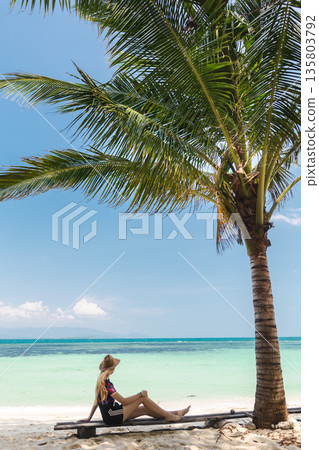 Young woman sitting under palm tree on tropical beach looking at turquoise sea and horizon, peaceful summer travel moment with clear sky Young woman sitting under palm tree on tropical beach looking at turquoise sea and horizon, peaceful summer travel moment with clear sky 135803792