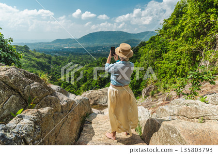 Young woman taking smartphone photo of lush tropical valley and mountain landscape from rocky waterfall viewpoint on Koh Samui Thailand with copy space 135803793