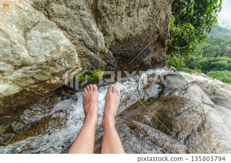 Bare feet above flowing waterfall on smooth rock ledge in tropical jungle, relaxing travel moment with fresh mountain stream and green forest background Bare feet above flowing waterfall on smooth rock ledge in tropical jungle, relaxing travel moment with fresh mountain stream and green forest background 135803794