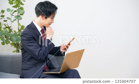 A male business person thinking while looking at a laptop and smartphone in the office A male business person thinking while looking at a laptop and smartphone in the office 135804707