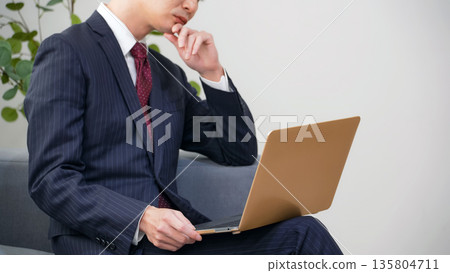 辦公室裡，一位男性商務人士一邊看著筆記型電腦一邊思考。 135804711