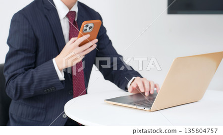 Male business person using laptop and smartphone in office Male business person using laptop and smartphone in office 135804757