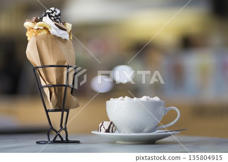 Ice cream dessert in wafer cup with chocolate cookies and and mug of coffee with marshmallows on porcelain plate on blurred colorful copy space background. Ice cream dessert in wafer cup with chocolate cookies and and mug of coffee with marshmallows on porcelain plate on blurred colorful copy space background. 135804915