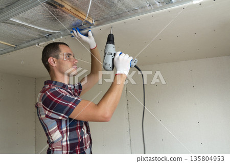 Young man in goggles fixing drywall suspended ceiling to metal frame using electrical screwdriver on ceiling insulated with shiny aluminum foil. Renovation, construction, do it yourself concept. Young man in goggles fixing drywall suspended ceiling to metal frame using electrical screwdriver on ceiling insulated with shiny aluminum foil. Renovation, construction, do it yourself concept. 135804953