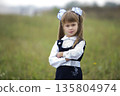 Close-up portrait of cute adorable confident first grader girl in school uniform and white bows in long blond hair on blurred light green sunny outdoors copy space background. 135804974