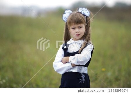 Close-up portrait of cute adorable confident first grader girl in school uniform and white bows in long blond hair on blurred light green sunny outdoors copy space background. Close-up portrait of cute adorable confident first grader girl in school uniform and white bows in long blond hair on blurred light green sunny outdoors copy space background. 135804974