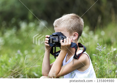 Profile close-up portrait of young blond cute handsome child boy with camera taking pictures outdoors on bright sunny spring or summer day on blurred light green grassy copy space background. Profile close-up portrait of young blond cute handsome child boy with camera taking pictures outdoors on bright sunny spring or summer day on blurred light green grassy copy space background. 135804980