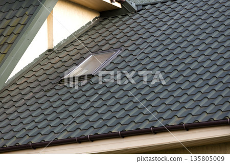Close-up detail of new small attic plastic window installed in dark green shingled house roof background. Professionally done building and construction work, roofing and installation concept. Close-up detail of new small attic plastic window installed in dark green shingled house roof background. Professionally done building and construction work, roofing and installation concept. 135805009