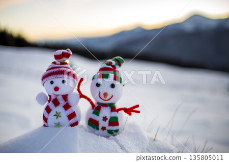 Two small funny toys baby snowman in knitted hats and scarves in deep snow outdoors on blurred mountains landscape background. Happy New Year and Merry Christmas greeting card theme. 135805011