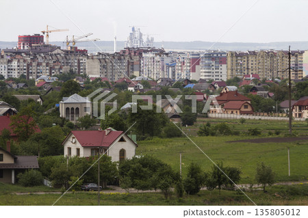 Wide beautiful summer panorama of quiet suburban area with nice comfortable cottages and green gardens on background of modern developing city with high buildings and working constructions cranes. 135805012