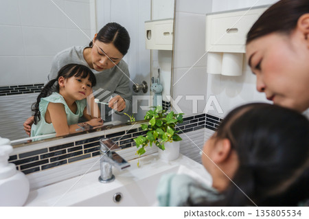 stubborn child girl refusing to brush her teeth in the bathroom. mother is trying to encourage her unhappy daughter during the morning routine, while child looks grumpy and resistant. stubborn child girl refusing to brush her teeth in the bathroom. mother is trying to encourage her unhappy daughter during the morning routine, while child looks grumpy and resistant. 135805534