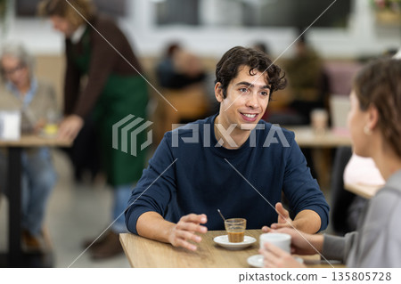 Happy young guy and pretty girl on date over cup of coffee at table in light cafe, face of guy and back of girl Happy young guy and pretty girl on date over cup of coffee at table in light cafe, face of guy and back of girl 135805728
