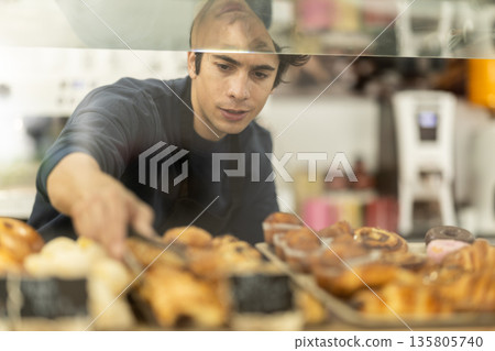 Male seller putting fresh pastries on plate in cafe bakery Male seller putting fresh pastries on plate in cafe bakery 135805740