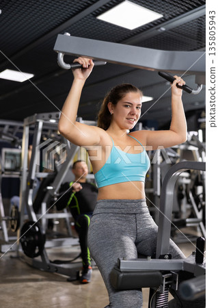 Active young woman training at shoulder press machine in gym Active young woman training at shoulder press machine in gym 135805943