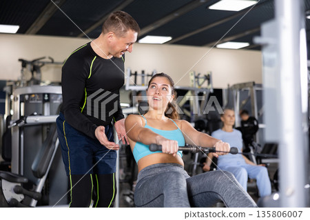 Male fitness trainer is engaged explains how to exercise on the simulator to an elderly woman in gym 135806007