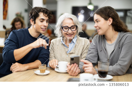 Positive elderly woman meeting and chatting with family and young couple showing photos on phone in cozy cafeteria 135806013