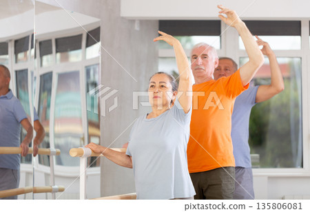 Ordinary elderly people stand in third position near ballet barre during group training in dance studio Ordinary elderly people stand in third position near ballet barre during group training in dance studio 135806081