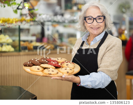 Elderly female waitress in apron invites to coffee shop and holding tray of fresh, delicious baked goods in hands Elderly female waitress in apron invites to coffee shop and holding tray of fresh, delicious baked goods in hands 135806123