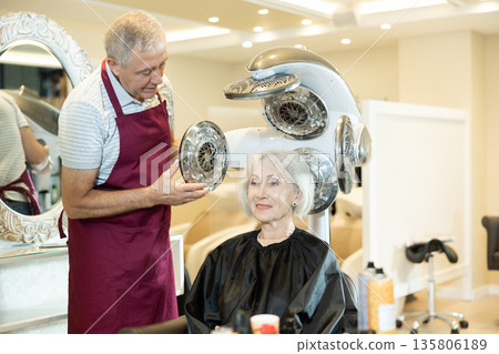 Apparatus for drying hair and creating a voluminous hairstyle. Professional male hairdresser makes a modern hairstyle for an mature woman Apparatus for drying hair and creating a voluminous hairstyle. Professional male hairdresser makes a modern hairstyle for an mature woman 135806189