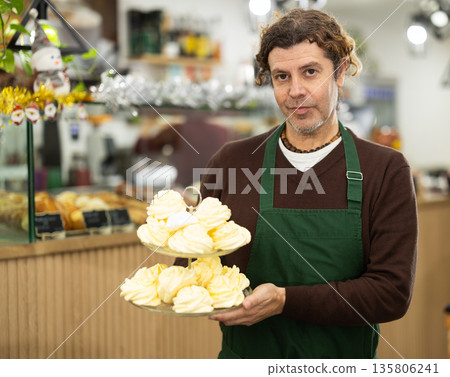 Handsome middle-aged male waiter in apron invites to coffee shop and holding tray of fresh, delicious baked goods in hands Handsome middle-aged male waiter in apron invites to coffee shop and holding tray of fresh, delicious baked goods in hands 135806241