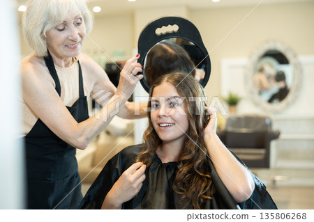Elderly woman hairdresser holds mirror behind girl client and shows result of work styling Elderly woman hairdresser holds mirror behind girl client and shows result of work styling 135806268