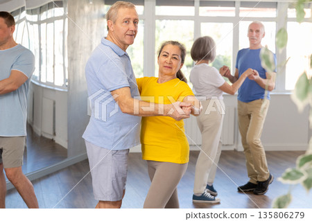 Positive senior woman and man practicing tango dance moves as a couple during a group celebration in dance studio Positive senior woman and man practicing tango dance moves as a couple during a group celebration in dance studio 135806279