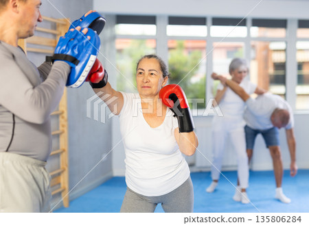 Senior lady and male couch are boxing in gym, man helps student to work out force of blow. Senior lady and male couch are boxing in gym, man helps student to work out force of blow. 135806284