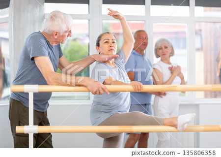 Professional senior male ballet trainer teaching group of dancers, helps to coordinate movements correctly to mature woman near ballet barre in dancing hall Professional senior male ballet trainer teaching group of dancers, helps to coordinate movements correctly to mature woman near ballet barre in dancing hall 135806358
