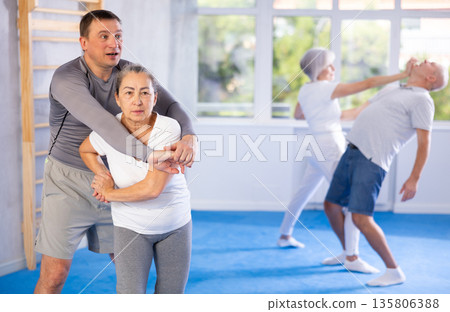 Trainer and mature woman train painful hold on self-defense course in sport gym Trainer and mature woman train painful hold on self-defense course in sport gym 135806388
