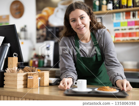 Female barista is standing near the counter in a coffee shop and preparing an order Female barista is standing near the counter in a coffee shop and preparing an order 135806398