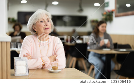 Elderly woman sitting at a table in a coffee shop with a cup of aromatic coffee Elderly woman sitting at a table in a coffee shop with a cup of aromatic coffee 135806416